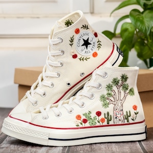 May include: A pair of white high-top Converse sneakers with red stitching. The sneakers are embroidered with floral designs and a cactus motif. The left shoe has the Converse logo and the text "Chuck Taylor All Star".