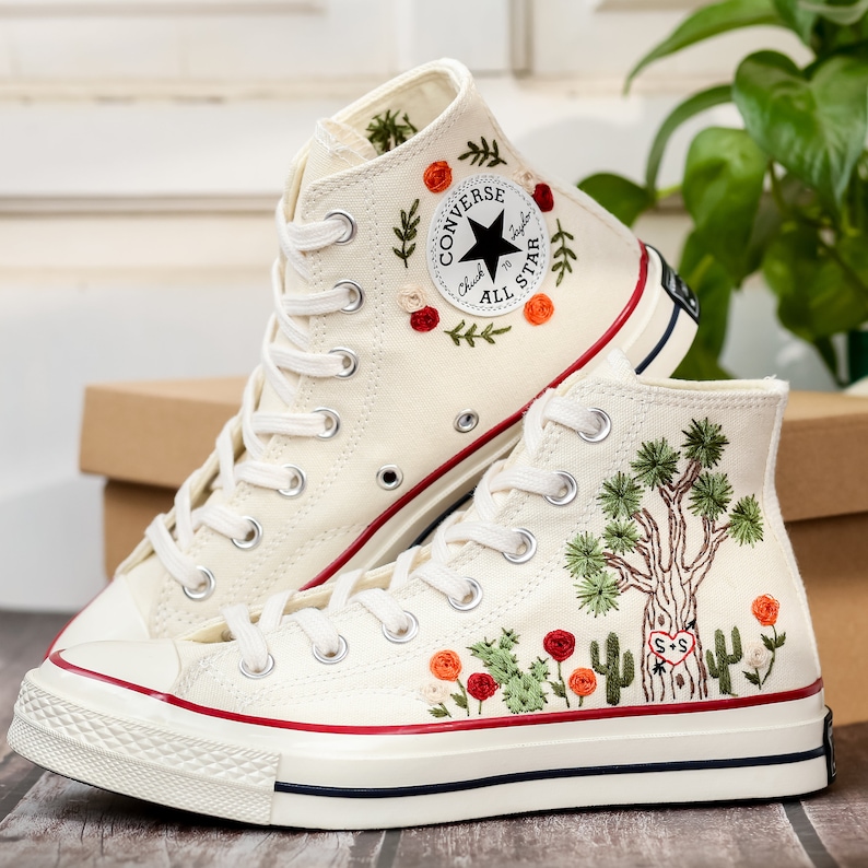 May include: A pair of white high-top Converse sneakers with red stitching. The sneakers are embroidered with floral designs and a cactus motif. The left shoe has the Converse logo and the text "Chuck Taylor All Star".