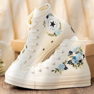 May include: Cream-colored high-top sneakers with floral embroidery. The sneakers feature blue and green embroidered flowers and leaves. The Converse All Star logo is visible on the side. The shoes have white laces and a rubber sole.