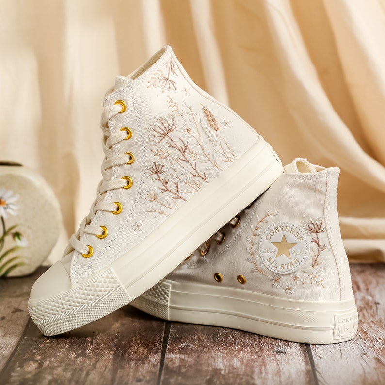 May include: A pair of white high-top canvas trainers with gold eyelets and embroidered floral details. The trainers feature a white rubber sole and the Converse All Star logo on the side.
