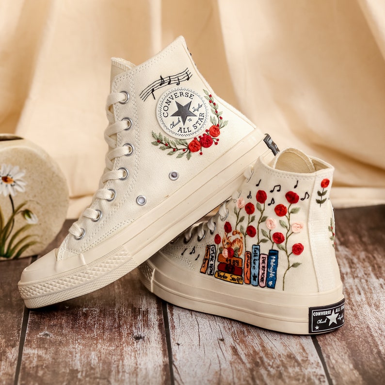 May include: A pair of white high-top canvas sneakers with embroidered floral and musical note designs. One shoe features a red rose and musical notes, while the other has a row of colorful books and red roses.
