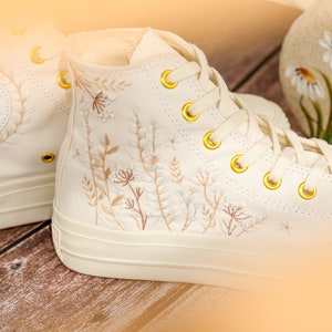 May include: A pair of white high-top Converse sneakers with gold eyelets and delicate floral embroidery. The embroidery features a variety of small flowers and leaves in shades of beige and brown.