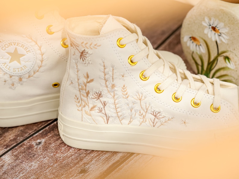 May include: A pair of white high-top Converse sneakers with gold eyelets and delicate floral embroidery. The embroidery features a variety of small flowers and leaves in shades of beige and brown.