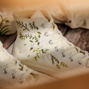May include: A pair of off-white canvas high-top sneakers with white laces. The sneakers are embroidered with delicate floral designs in shades of green and white.