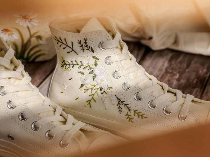 May include: A pair of off-white canvas high-top sneakers with white laces. The sneakers are embroidered with delicate floral designs in shades of green and white.