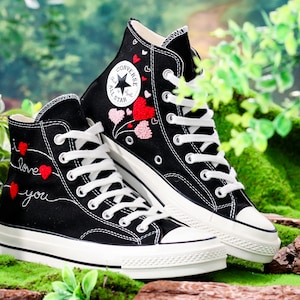 Op de afbeelding: Een paar zwarte Converse sneakers met hoge schacht, witte veters en een witte zool. De sneakers zijn versierd met rode en roze geborduurde harten en de woorden "Ik hou van je".
