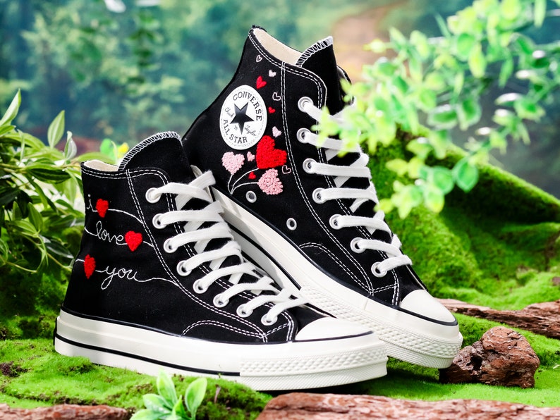 Op de afbeelding: Een paar zwarte Converse sneakers met hoge schacht, witte veters en een witte zool. De sneakers zijn versierd met rode en roze geborduurde harten en de woorden "Ik hou van je".