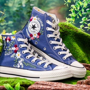 May include: A pair of blue high-top Converse trainers with white laces and a white rubber sole. The trainers are embroidered with colourful flowers and leaves.