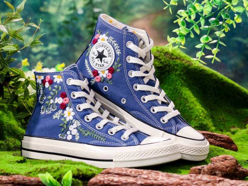 May include: A pair of blue high-top Converse trainers with white laces and a white rubber sole. The trainers are embroidered with colourful flowers and leaves.