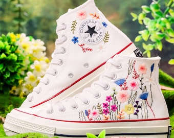 Può includere: Sneakers bianche in tela alta con ricami floreali e il logo Converse. Le sneakers hanno una suola in gomma bianca e una striscia rossa attorno al bordo.