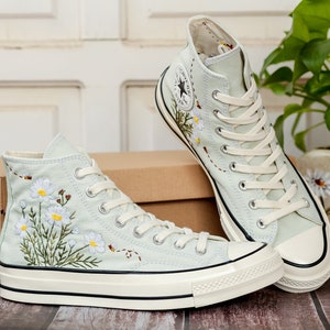 May include: A pair of light blue high-top canvas sneakers with white soles and white laces. The sneakers are embroidered with a floral design of white daisies and green leaves. The sneakers are sitting on a wooden surface.