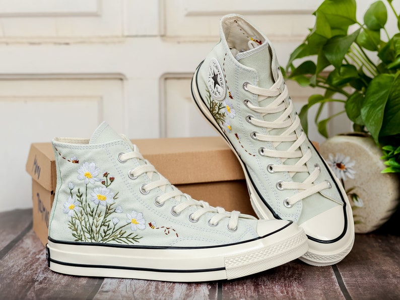 May include: A pair of light blue high-top canvas sneakers with white soles and white laces. The sneakers are embroidered with a floral design of white daisies and green leaves. The sneakers are sitting on a wooden surface.