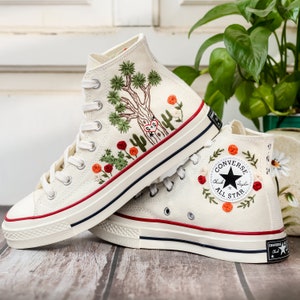 May include: A pair of white high-top Converse sneakers with embroidered floral designs and a cactus. The left shoe has a large cactus and the right shoe has the Converse logo with a star.