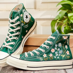Puede incluir: Un par de zapatillas altas de lona verde con cordones y suelas blancas. Las zapatillas est&aacute;n adornadas con dise&ntilde;os florales bordados en blanco, rosa y azul. El logotipo de Converse All Star es visible.