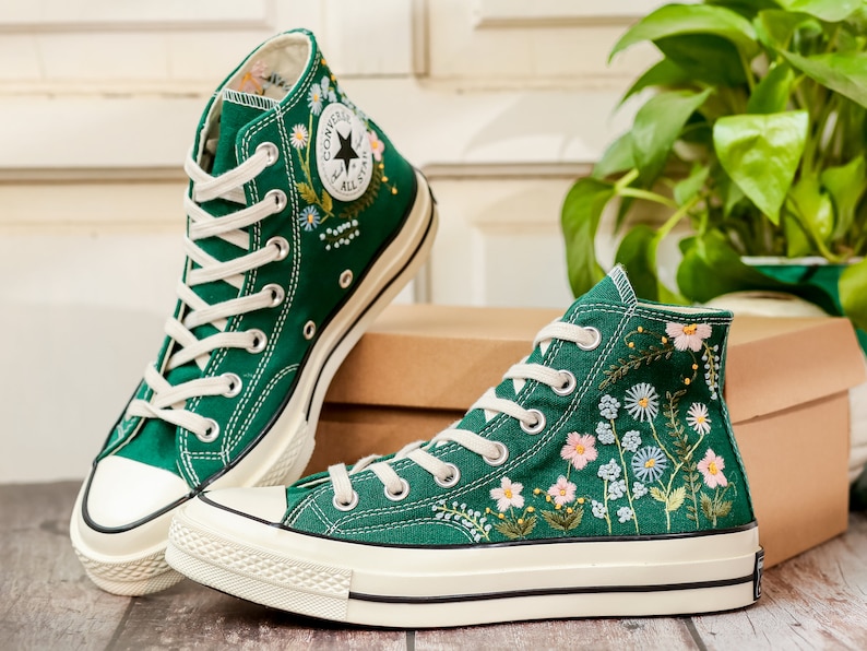 Puede incluir: Un par de zapatillas altas de lona verde con cordones y suelas blancas. Las zapatillas est&aacute;n adornadas con dise&ntilde;os florales bordados en blanco, rosa y azul. El logotipo de Converse All Star es visible.
