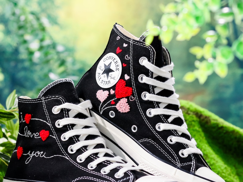 Op de afbeelding: Een paar zwarte Converse sneakers met hoge schacht en witte veters. De ene schoen heeft een rood en roze hart geborduurd ontwerp, terwijl de andere de woorden "Ik hou van je" in rood en wit geborduurd heeft.