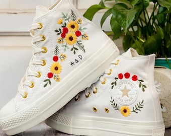 May include: White high-top sneakers with embroidered sunflowers, red berries, and green leaves. The sneakers have gold eyelets and the text "A & D" on the side. The Converse logo is visible on the side of the other shoe.