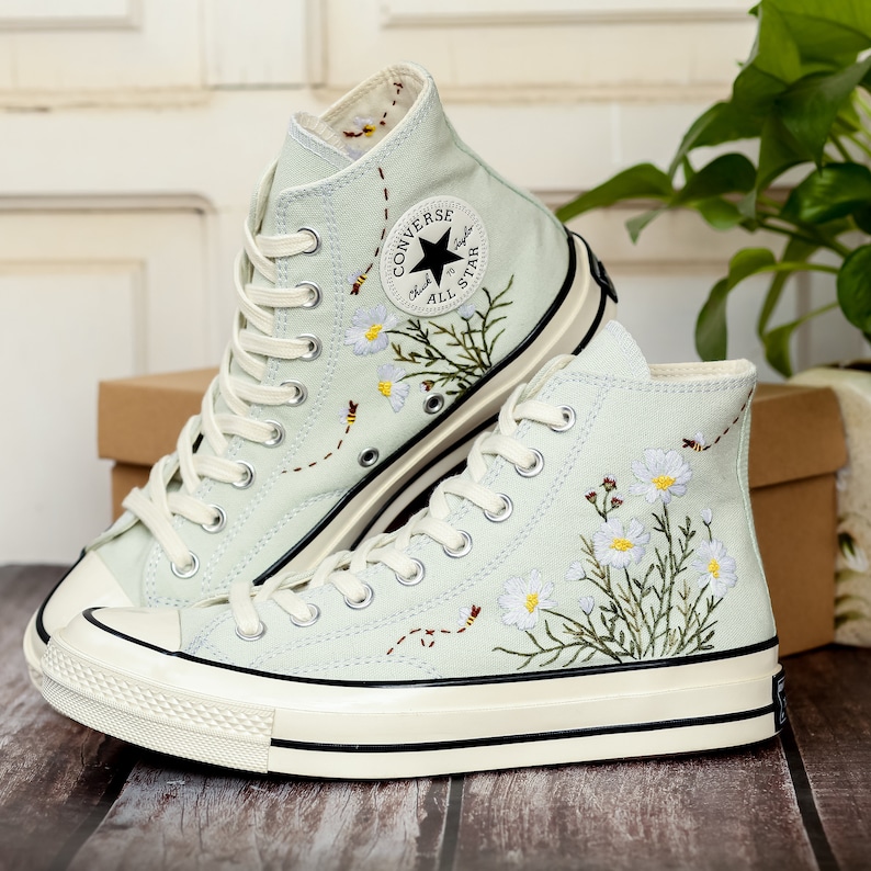 May include: A pair of light blue high-top Converse sneakers with white daisies and bees embroidered on the canvas. The sneakers have a white rubber sole and a black Converse All Star logo on the side.
