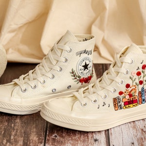 May include: A pair of off-white high-top Converse sneakers with embroidered floral designs and musical notes. One shoe features a red rose and the text "Converse All Star", while the other shoe features a stack of books with titles like "Speak Now" and "Red".