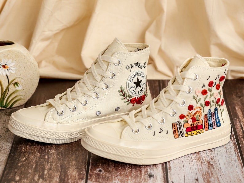 May include: A pair of off-white high-top Converse sneakers with embroidered floral designs and musical notes. One shoe features a red rose and the text "Converse All Star", while the other shoe features a stack of books with titles like "Speak Now" and "Red".