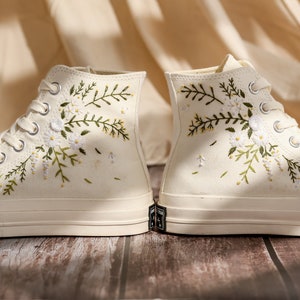 May include: A pair of white high-top canvas sneakers with embroidered floral designs. The flowers are white and green and the sneakers have white laces.