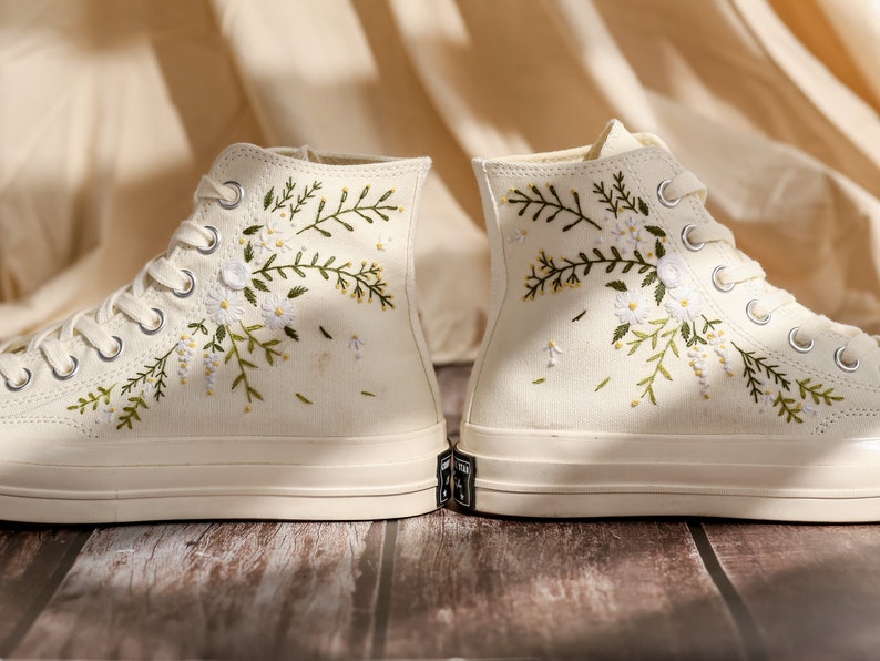 May include: A pair of white high-top canvas sneakers with embroidered floral designs. The flowers are white and green and the sneakers have white laces.