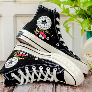 May include: A pair of black high-top Converse sneakers with white soles and laces. The sneakers are embroidered with a floral design and red mushrooms. The Converse All Star logo is visible on the side of the shoe.