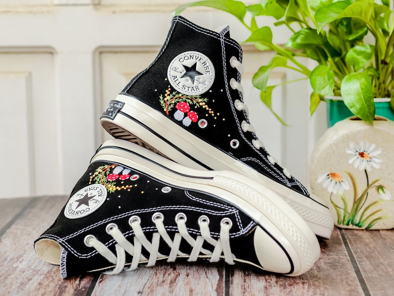 May include: A pair of black high-top Converse sneakers with white soles and laces. The sneakers are embroidered with a floral design and red mushrooms. The Converse All Star logo is visible on the side of the shoe.