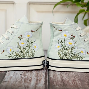 May include: A pair of light blue high-top sneakers with white embroidered flowers and green leaves. The sneakers have a white rubber sole with a black trim.