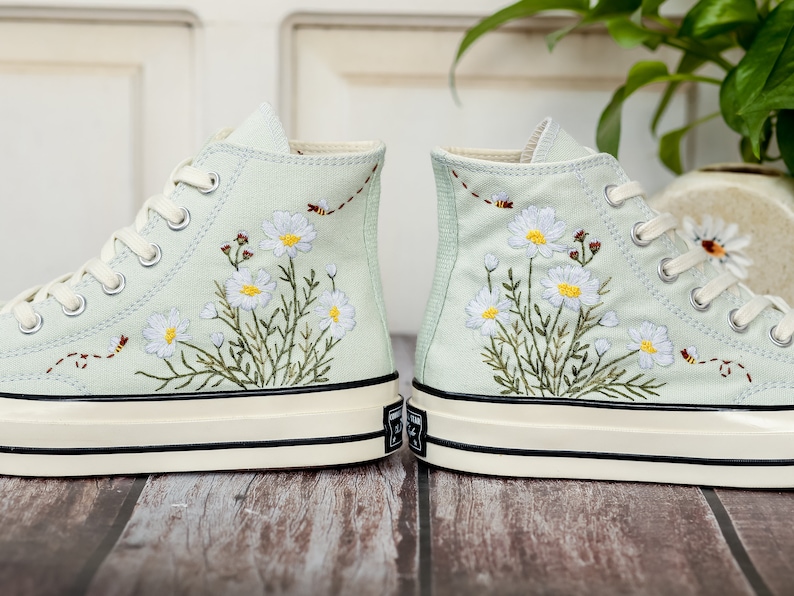 May include: A pair of light blue high-top sneakers with white embroidered flowers and green leaves. The sneakers have a white rubber sole with a black trim.
