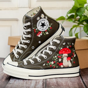 May include: A pair of olive green high-top Converse trainers with white soles. The trainers are embroidered with red mushrooms, white daisies, and green leaves. The Converse logo is visible on the side of the trainers.