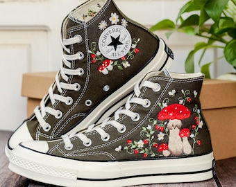 May include: A pair of olive green high-top Converse sneakers with white soles. The sneakers are embroidered with red mushrooms, white daisies, and green leaves. The Converse logo is visible on the side of the sneakers.