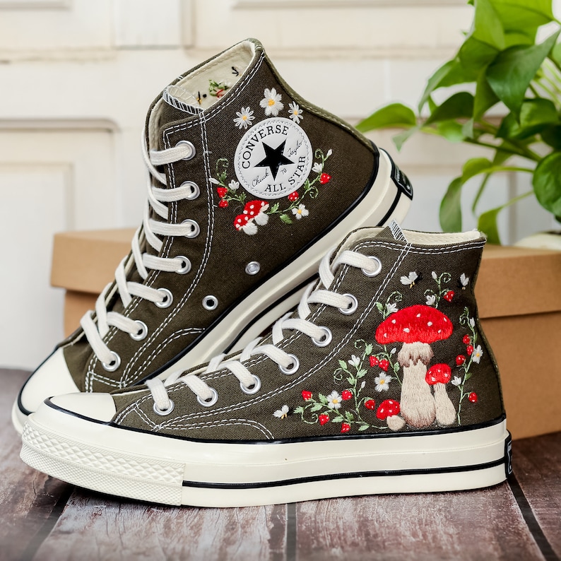 May include: A pair of olive green high-top Converse trainers with white soles. The trainers are embroidered with red mushrooms, white daisies, and green leaves. The Converse logo is visible on the side of the trainers.