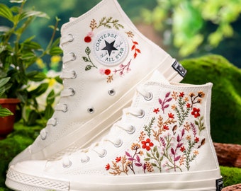 May include: White high-top Converse sneakers with floral embroidery. The sneakers feature a classic Converse design with a white canvas upper, black laces, and a white rubber sole. The floral embroidery is in shades of red, pink, green, and yellow.