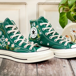 Puede incluir: Un par de zapatillas altas de lona verde esmeralda, adornadas con delicados bordados florales en blanco, rosa y amarillo. Las zapatillas tienen cordones blancos, una suela de goma color crema y el ic&oacute;nico logotipo de Converse All Star.