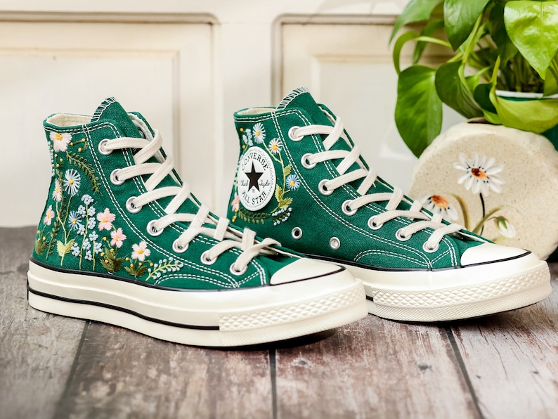 Puede incluir: Un par de zapatillas altas de lona verde esmeralda, adornadas con delicados bordados florales en blanco, rosa y amarillo. Las zapatillas tienen cordones blancos, una suela de goma color crema y el ic&oacute;nico logotipo de Converse All Star.