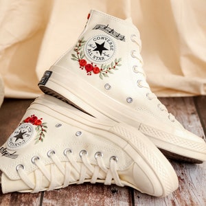May include: A pair of white high-top Converse sneakers with red floral embroidery and musical notes. The Converse All Star logo is visible on the side of the shoes.