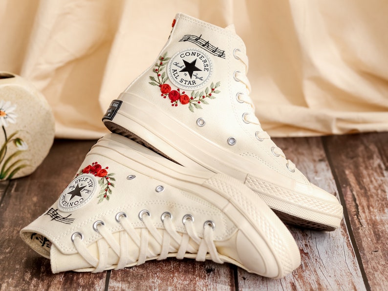 May include: A pair of white high-top Converse sneakers with red floral embroidery and musical notes. The Converse All Star logo is visible on the side of the shoes.
