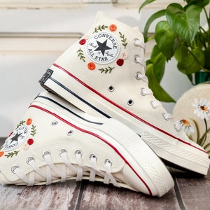 May include: A pair of white high-top Converse sneakers with red and green floral embroidery. The sneakers have a black and white Converse All Star logo on the side.