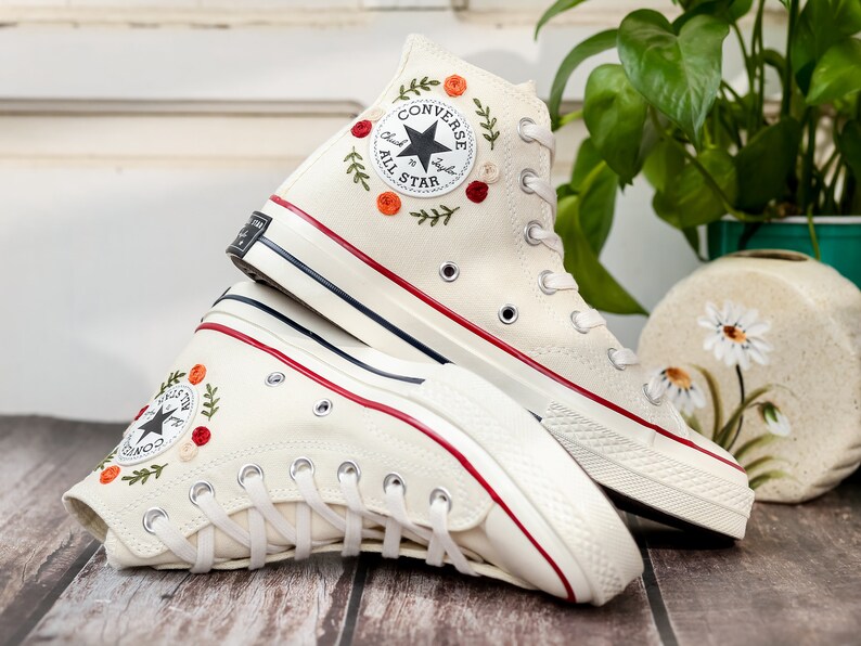 May include: A pair of white high-top Converse sneakers with red and green floral embroidery. The sneakers have a black and white Converse All Star logo on the side.