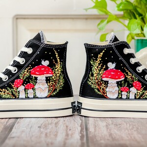 May include: A pair of black high-top canvas sneakers with white soles. The sneakers are decorated with embroidered red and white mushrooms, green leaves, and a snail.