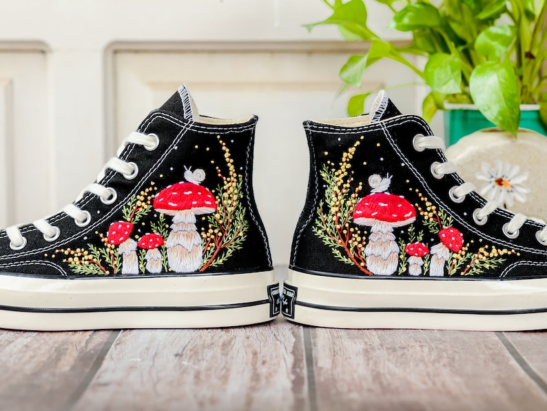 May include: A pair of black high-top canvas sneakers with white soles. The sneakers are decorated with embroidered red and white mushrooms, green leaves, and a snail.