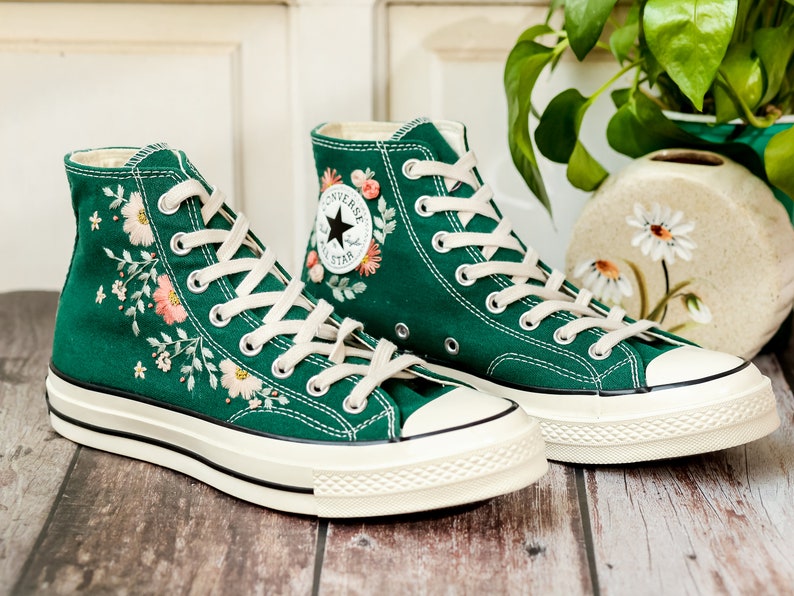 May include: A pair of green high-top canvas sneakers with white laces and a white rubber sole. The sneakers are embroidered with pink and white flowers and green leaves. The Converse All Star logo is visible on the side of the sneakers.