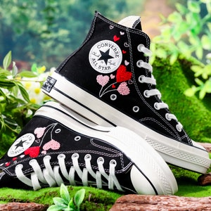 Op de afbeelding: Zwarte Converse sneakers met hoge schacht, witte zolen en roze en rode geborduurde harten. De sneakers hebben het Converse All Star logo aan de zijkant.
