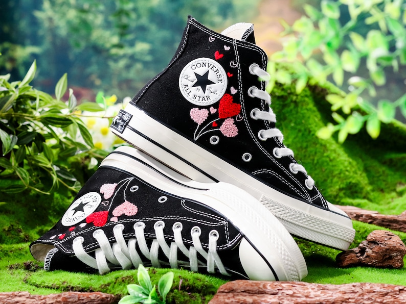 Op de afbeelding: Zwarte Converse sneakers met hoge schacht, witte zolen en roze en rode geborduurde harten. De sneakers hebben het Converse All Star logo aan de zijkant.