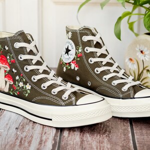 May include: A pair of olive green high-top canvas trainers with white laces and a white rubber sole. The trainers are embroidered with red and white mushrooms, flowers, and leaves.