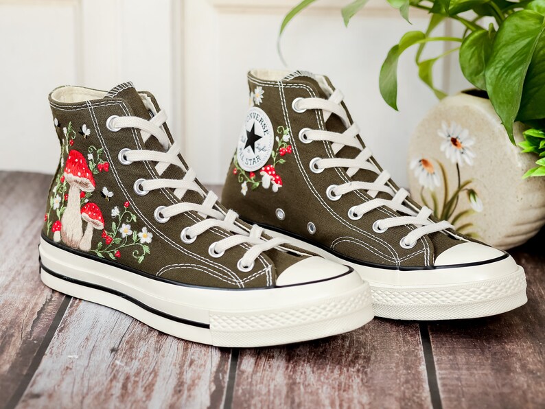 May include: A pair of olive green high-top canvas trainers with white laces and a white rubber sole. The trainers are embroidered with red and white mushrooms, flowers, and leaves.