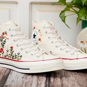May include: White high-top canvas sneakers with red, yellow and green embroidered flowers. The sneakers have a white rubber sole and a red stripe around the edge.