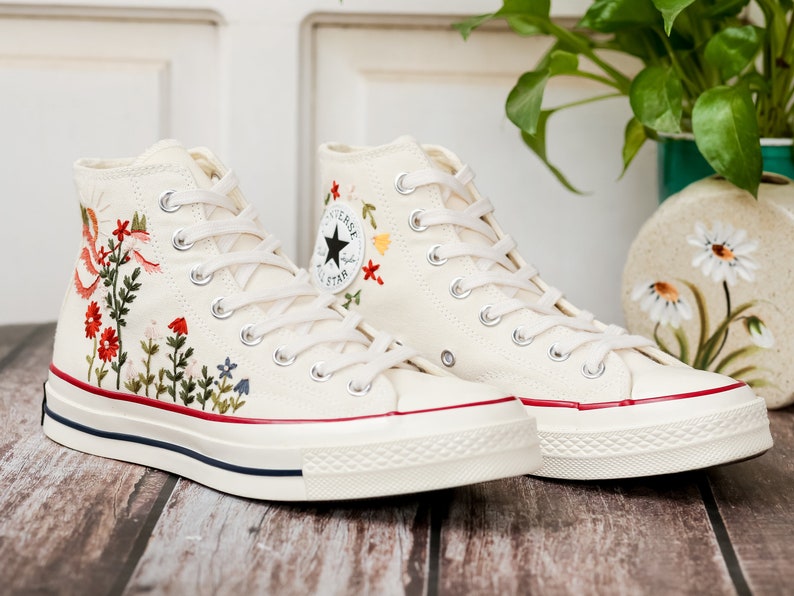 May include: White high-top canvas sneakers with red, yellow and green embroidered flowers. The sneakers have a white rubber sole and a red stripe around the edge.