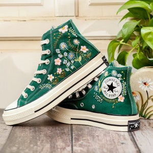 Puede incluir: Zapatillas altas verde esmeralda con bordados florales. Las zapatillas tienen cordones blancos, puntera de goma blanca y suela color crema. El logo de Converse All Star es visible en el lateral.
